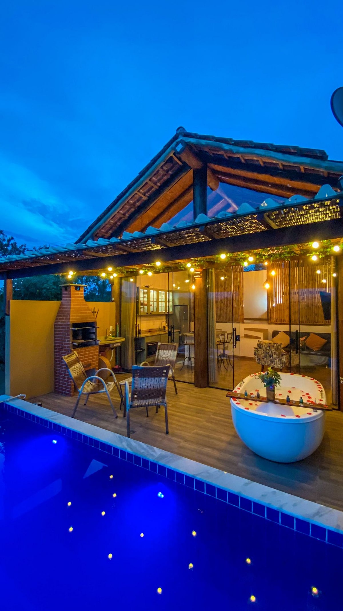An outdoor area features a wooden deck surrounding a private pool, illuminated by soft string lights. A cozy seating arrangement with two chairs sits nearby, alongside a grill and a stylish bathtub adorned with flower petals, offering a serene view of the evening sky.