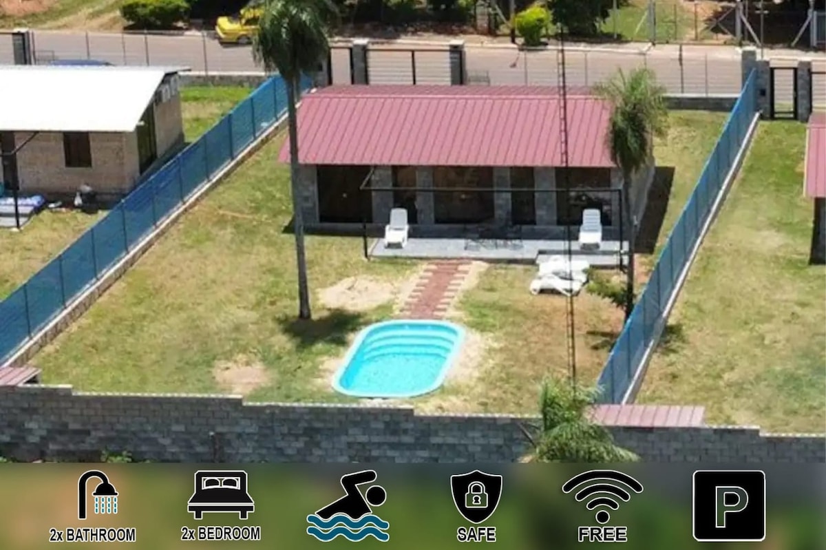 An aerial view of the property reveals a cozy bungalow surrounded by a spacious yard. A private pool is visible in the center, complemented by sun loungers. The house features a red roof and is enclosed by fencing, emphasizing a secure and private environment.