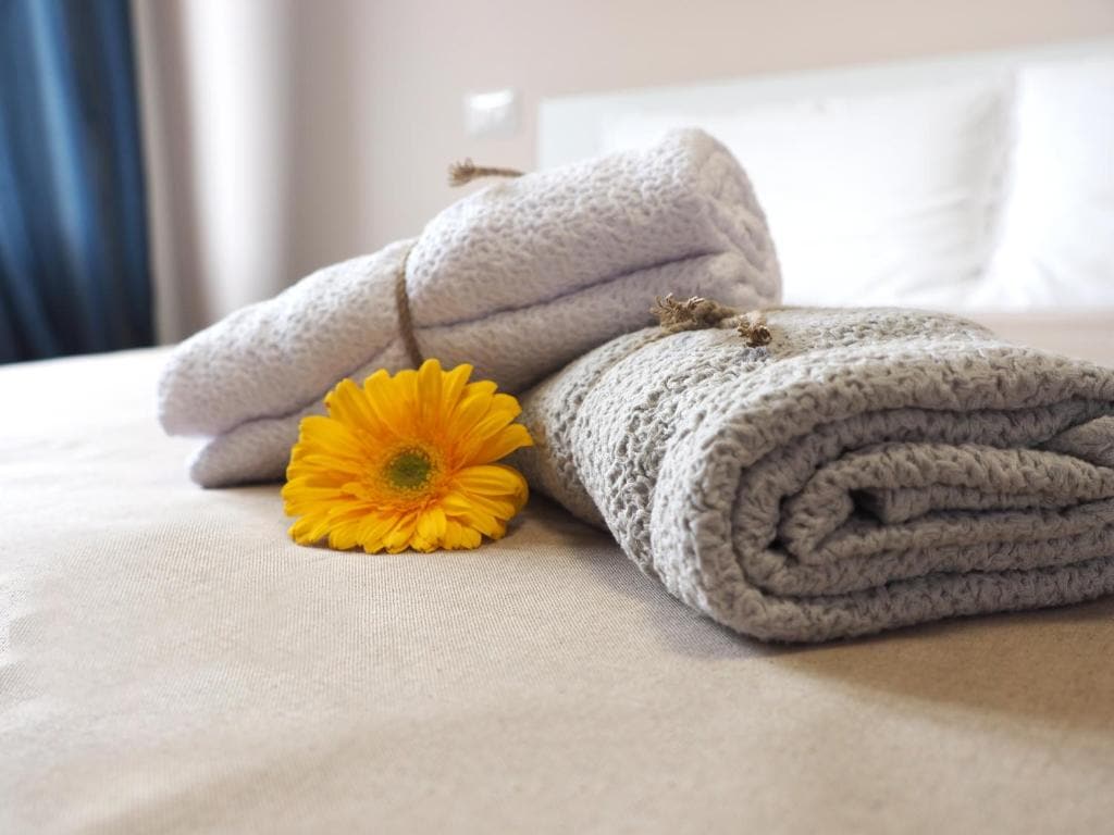 Fresh towels are neatly rolled and placed on a smooth, beige bedspread. A vibrant yellow flower adds a touch of color, providing a subtle accent to the inviting arrangement.