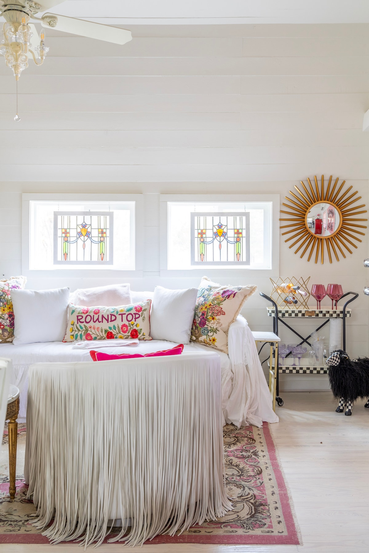 A charming living area features a cozy couch adorned with floral pillows and a decorative 'Round Top' pillow. Two stained glass windows allow natural light to illuminate the space. A round table with a fringe skirt complements a vintage-style bar cart, enhancing the inviting design.