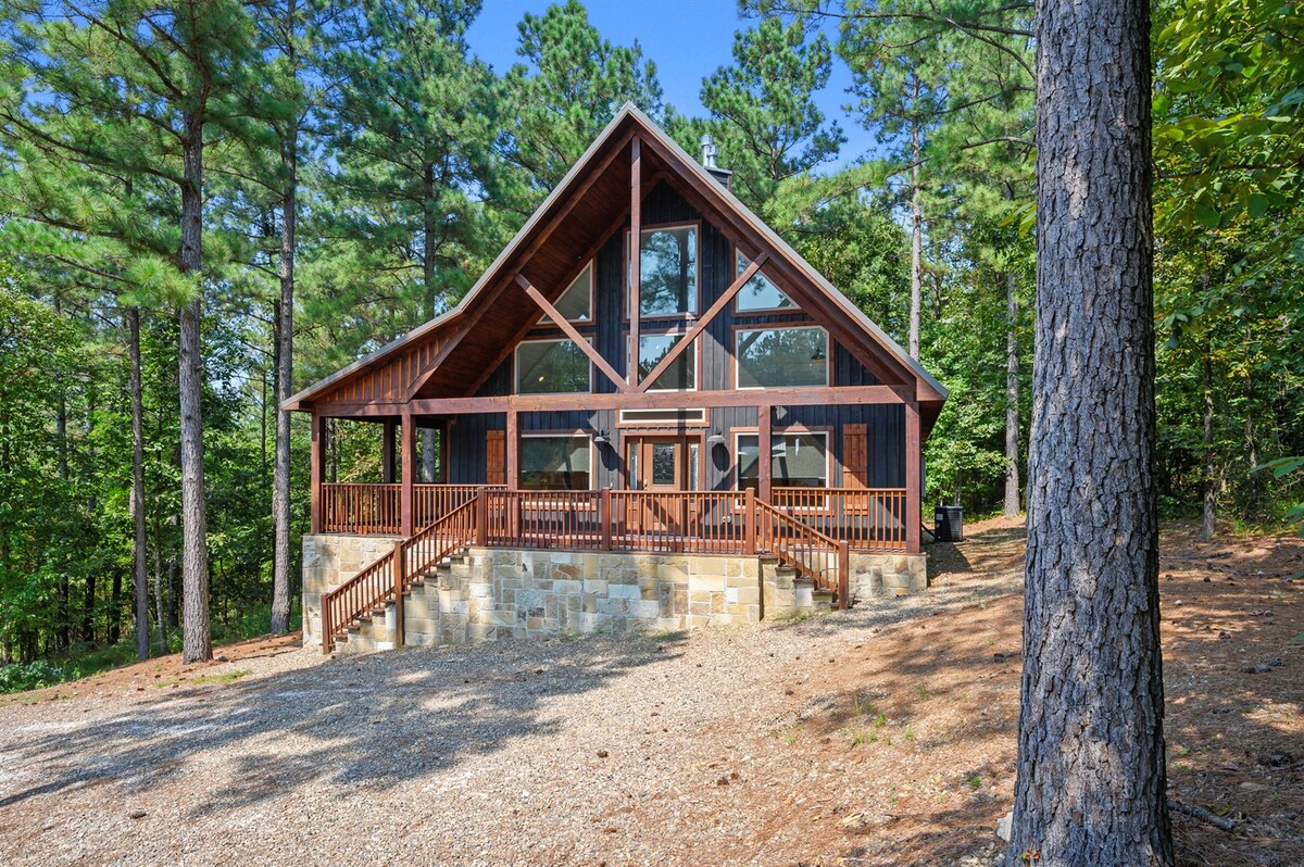 A charming cabin is surrounded by tall pine trees, displaying an inviting wooden facade with large windows. The exterior features a spacious front porch with a wooden railing, leading to outdoor steps. The setting offers a sense of tranquility and connection with nature.