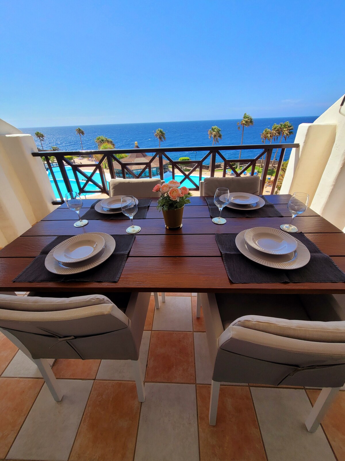 A dining table is set for four, featuring plates and wine glasses, with a view of the ocean and palm trees in the background. The space is framed by a balcony railing, allowing ample natural light to illuminate the area.