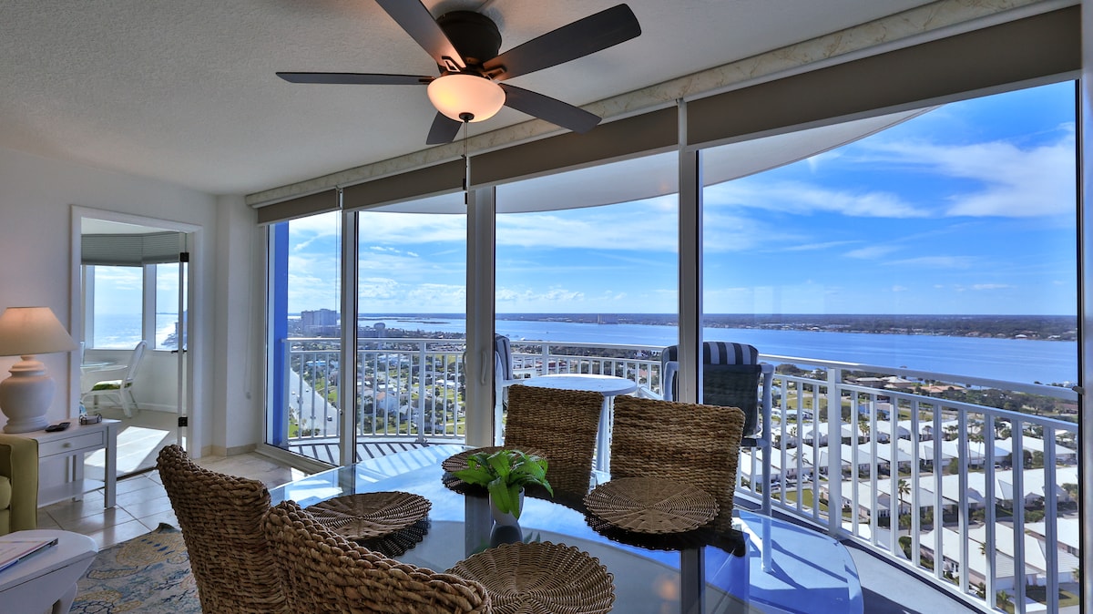 A modern dining area features glass table surrounded by woven chairs, with a view of the ocean through expansive glass sliding doors. Natural light enhances the space, while a ceiling fan adds a breezy sensation. Scenic vistas of the coastline and city are framed by the window.