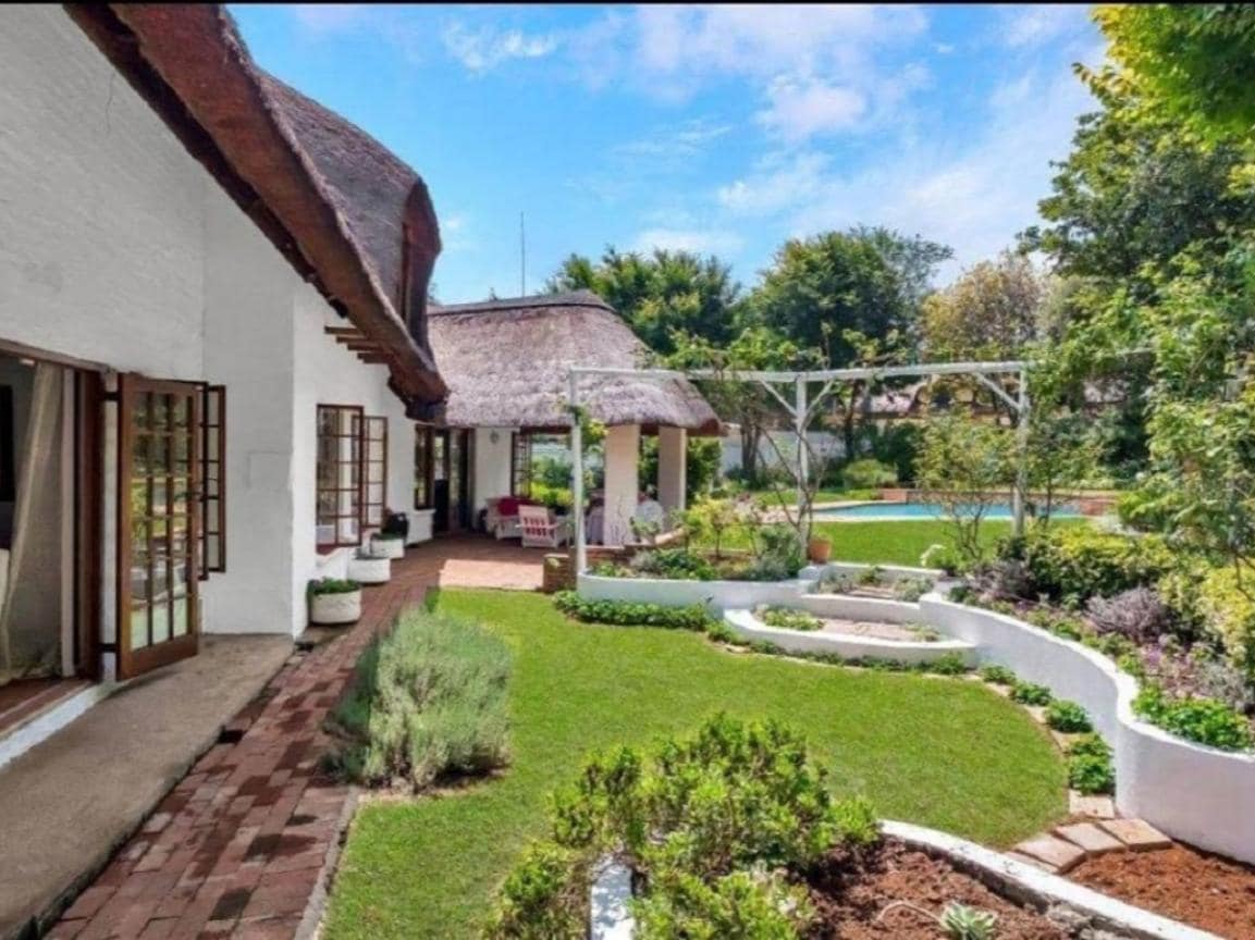 The exterior showcases a charming thatched roof structure surrounded by vibrant greenery and well-maintained gardens. A spacious lawn is complemented by flower beds and pathways, inviting leisurely strolls. A pool can be seen in the background, enhancing the outdoor atmosphere.