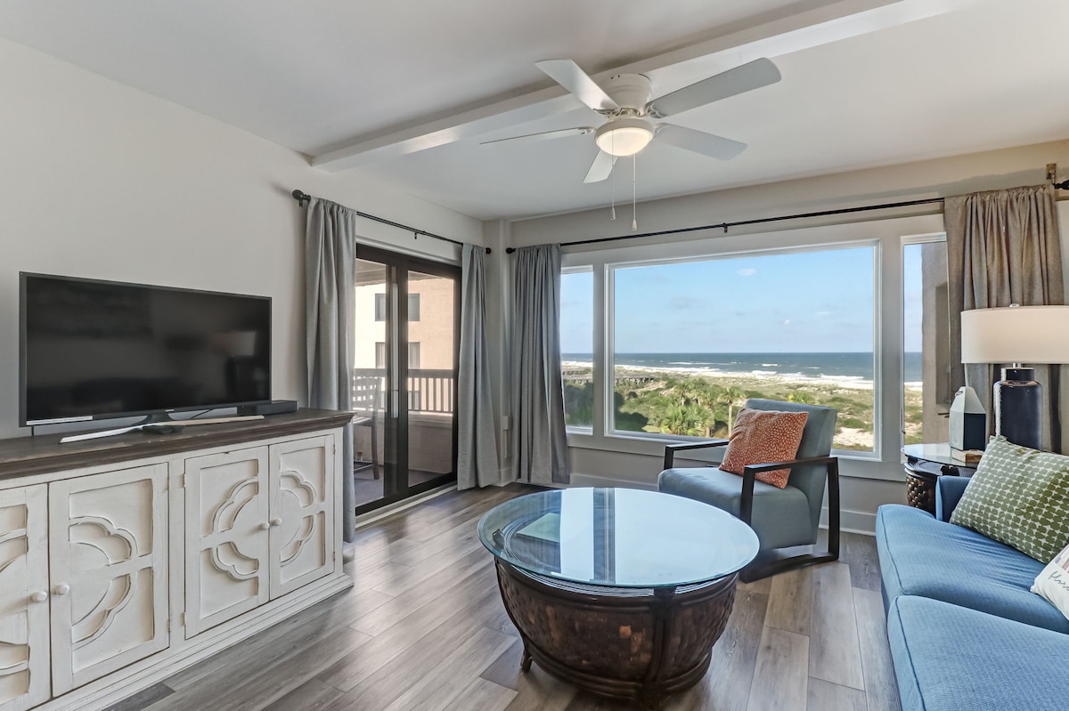 A spacious living area features large windows offering expansive ocean views. A comfortable sofa and an armchair are arranged around a circular coffee table. A television is positioned on the entertainment unit, with sliding glass doors leading to the private balcony.
