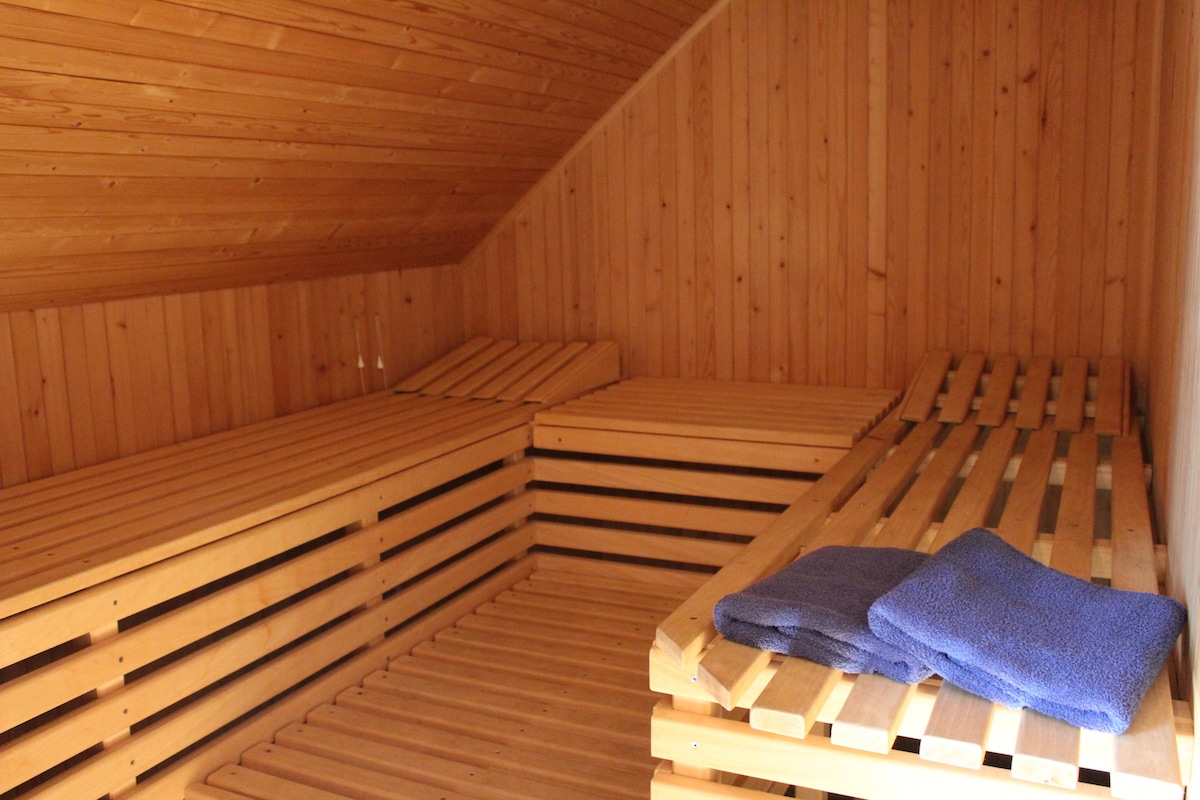 A wooden sauna interior is depicted, featuring slatted benches on three sides. Fresh towels are neatly placed on the benches, and the warm wood paneling enhances the cozy atmosphere. The ceiling is angled, contributing to the intimate design of the space.