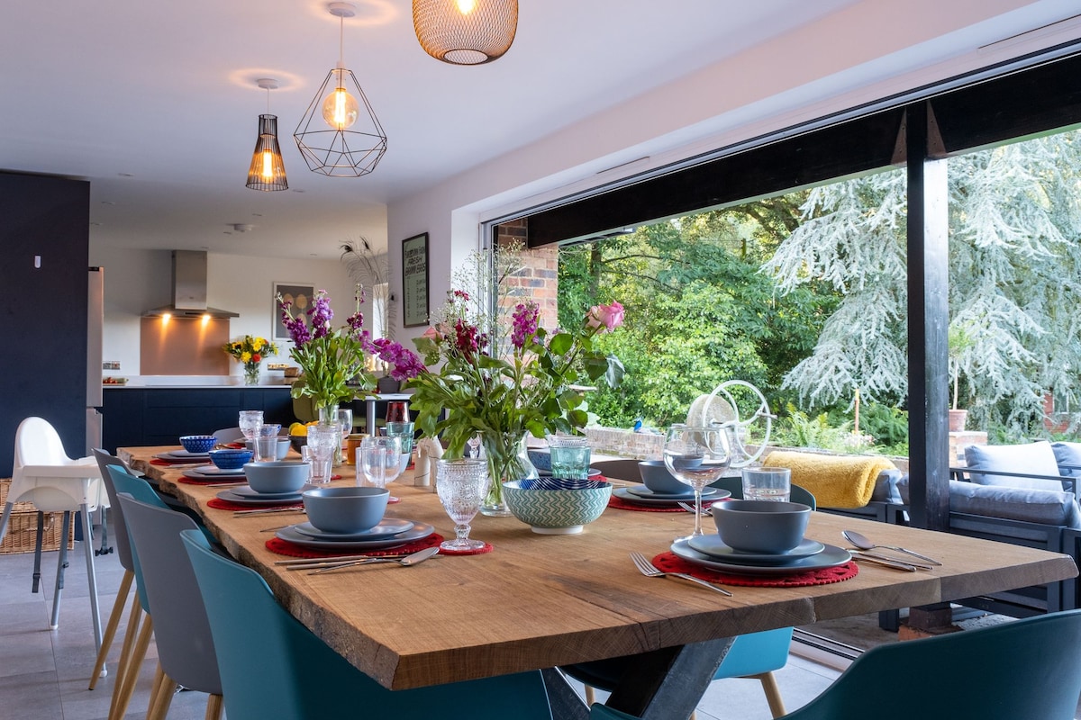 A large wooden dining table is set for a meal, featuring plates, glasses, and floral centerpieces. Bifold doors open to a lush garden, inviting natural light into the space. Modern lighting fixtures hang above, complementing the clean lines of the decor.