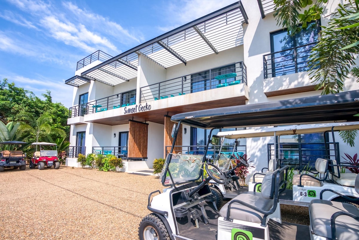 The exterior of Sunset Gecko Condos is shown, featuring a modern building design with multiple balconies. Golf carts are parked in front on a gravel driveway, surrounded by lush greenery and ornamental plants. Large windows provide a view of the vibrant outdoor space.