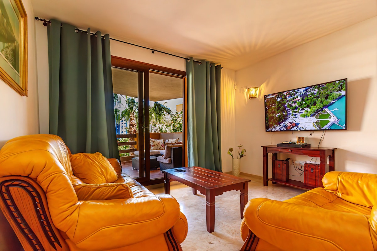 A cozy living area features two leather armchairs positioned around a wooden coffee table. A television is mounted on the wall opposite, and large glass doors lead to a terrace. Soft curtains frame the windows, allowing natural light to fill the space.