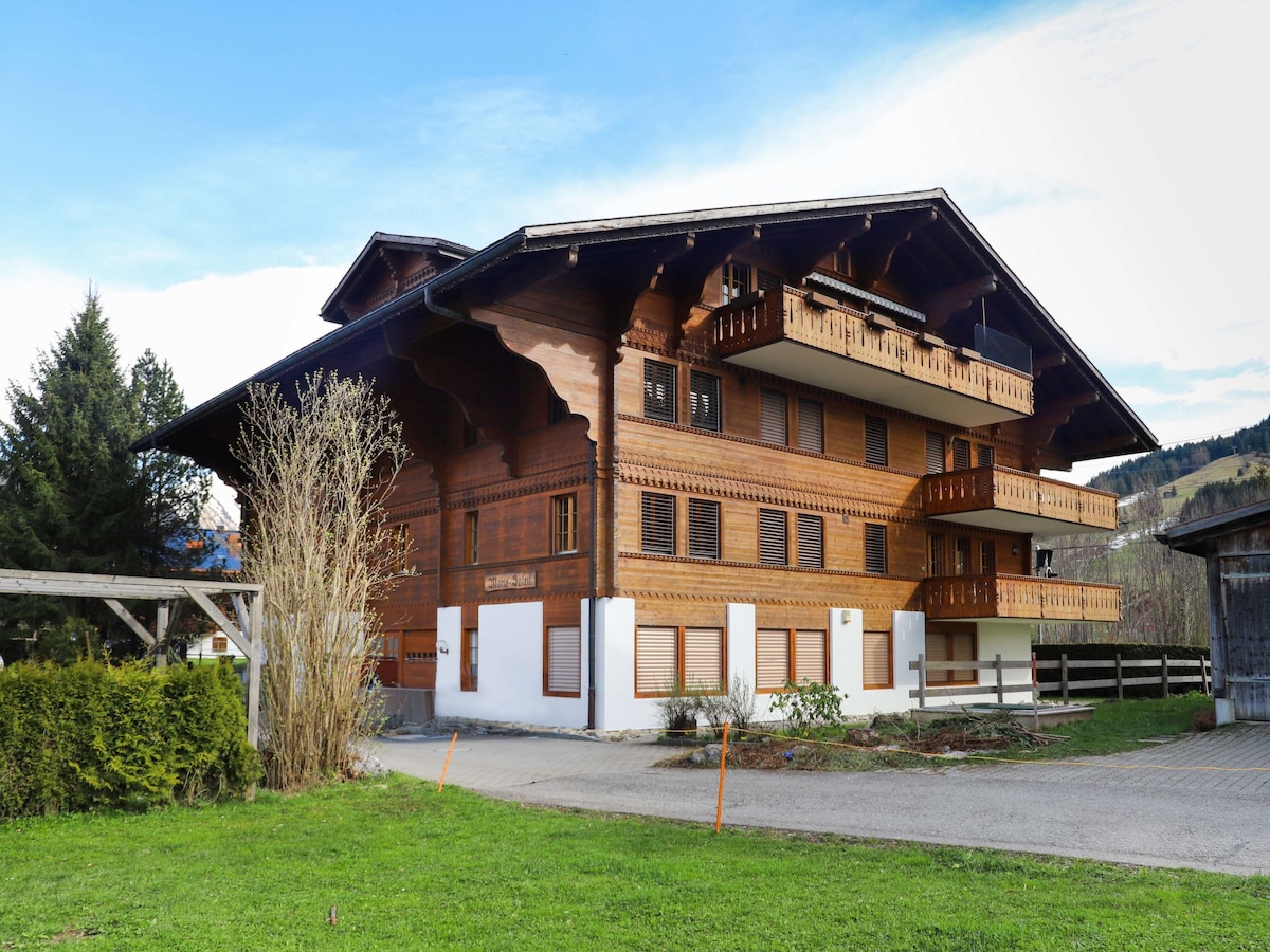 The building showcases a traditional Swiss chalet style with wooden balconies and decorative elements. Three floors are visible, with several windows allowing natural light in. Surrounding green space is complemented by well-maintained hedges and a gravel driveway, providing access to the entrance.