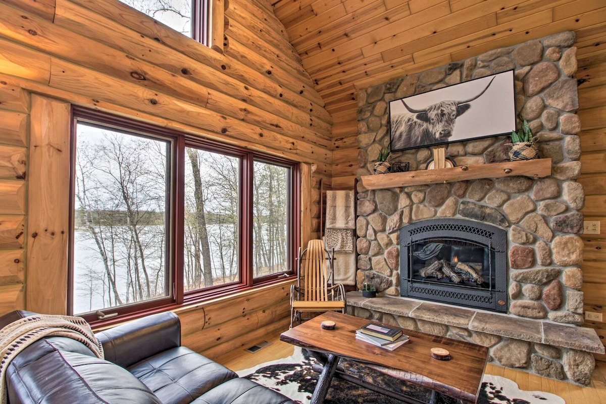 A cozy living room is highlighted by warm wooden walls and large windows offering views of Fawn Lake. A stone fireplace with a black mantel serves as a focal point, complemented by a wooden coffee table and a comfortable leather sofa. Natural light enhances the inviting atmosphere.