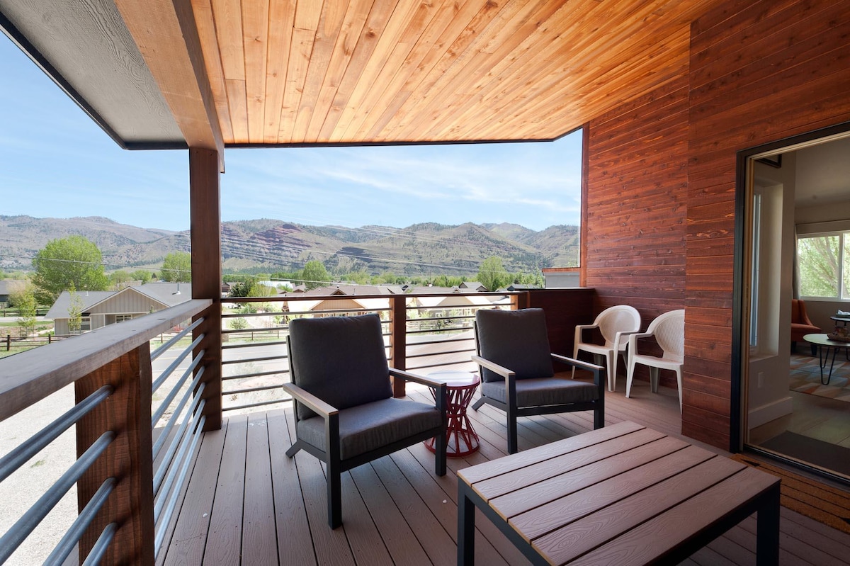 An outdoor balcony features comfortable seating with two chairs and a small table, offering views of the surrounding valley and red rock formations. The wood-paneled ceiling adds warmth, while the railing provides an open feel to the space.