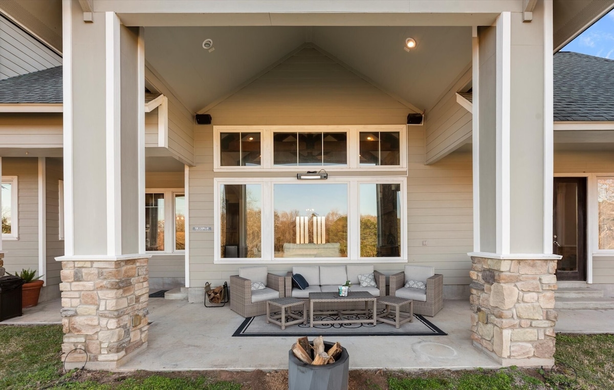 An outdoor seating area is situated under a covered porch, featuring a comfortable arrangement of gray wicker furniture and a low coffee table. Large glass windows provide views of the surrounding landscape, while a fire pit is located in the foreground.