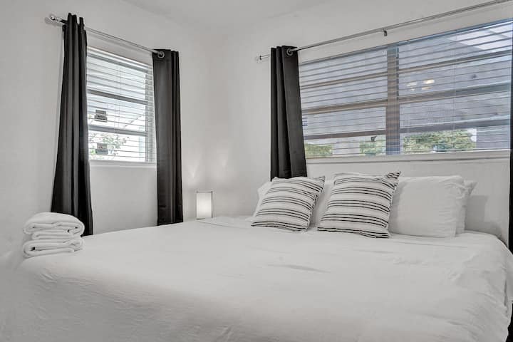 El dormitorio ofrece un ambiente acogedor con una lujosa cama, almohadas a rayas y decoración minimalista. La luz natural fluye a través de las grandes ventanas, acentuadas por elegantes cortinas negras. La iluminación suave crea un retiro relajante y acogedor.