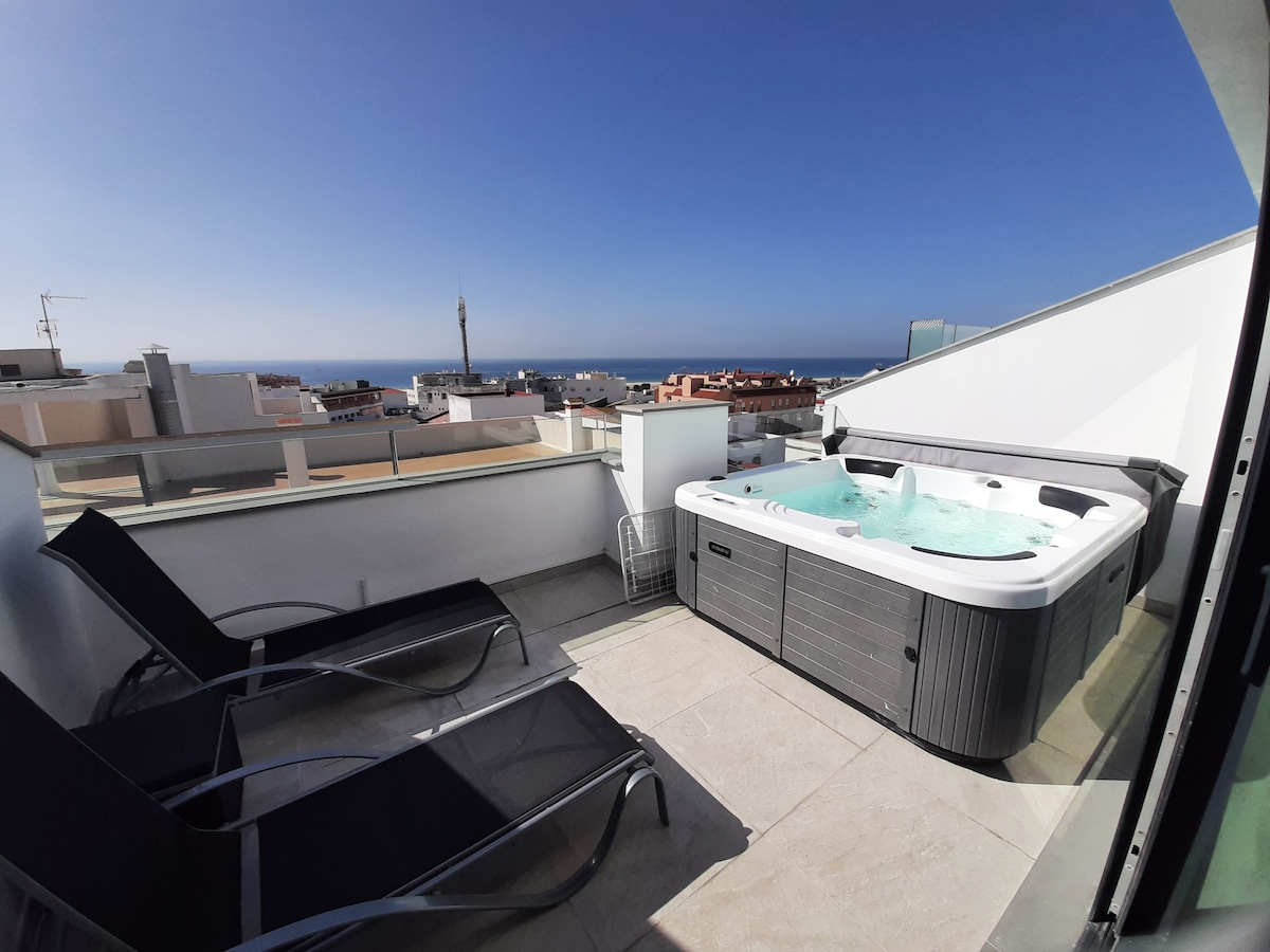 A private terrace offers a relaxing atmosphere, featuring a jacuzzi and two lounge chairs. The space is complemented by an unobstructed view of the ocean and the surrounding rooftops, with clear blue skies enhancing the outdoor experience.