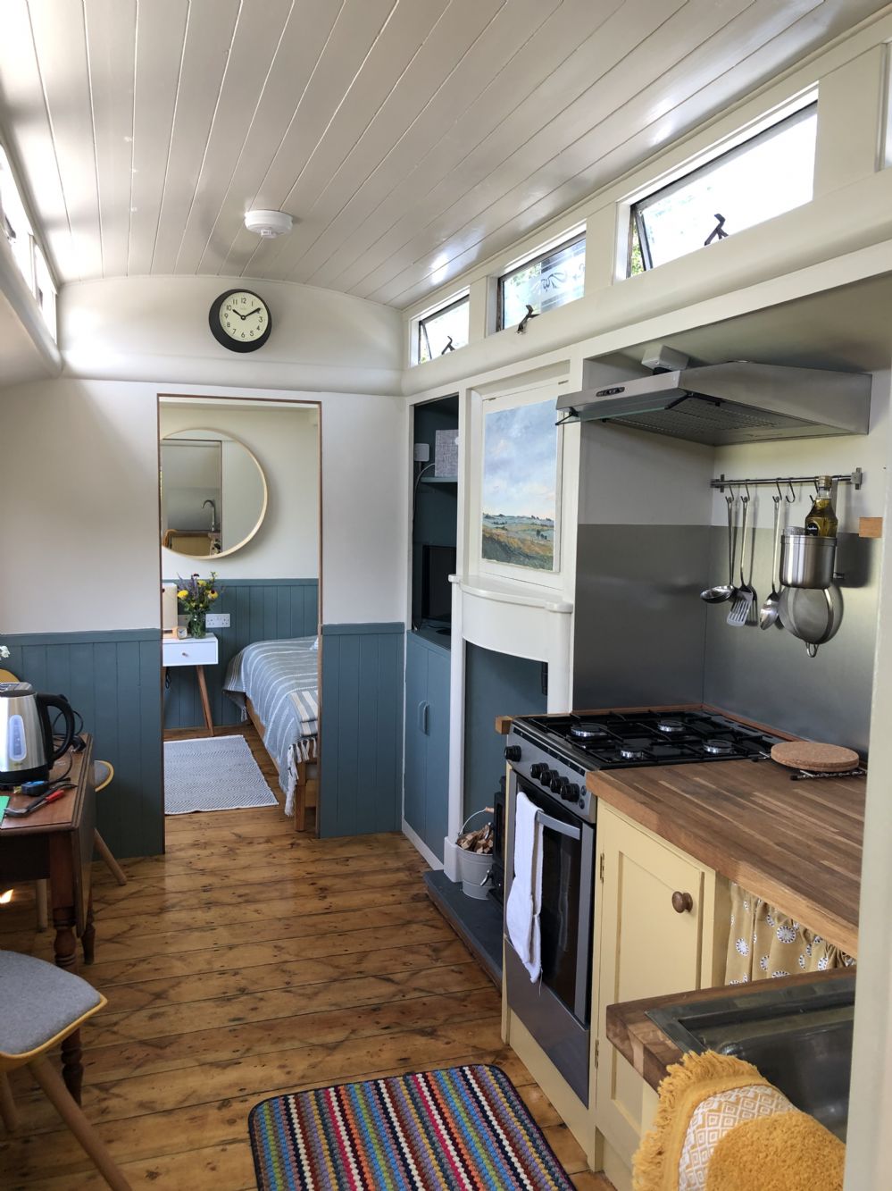 The interior of the showman’s wagon showcases a handcrafted kitchen area equipped with a stove and utensils. Natural light streams through original windows, illuminating the space. A cozy sleeping area is visible through an open doorway, featuring a round mirror and soft bedding.