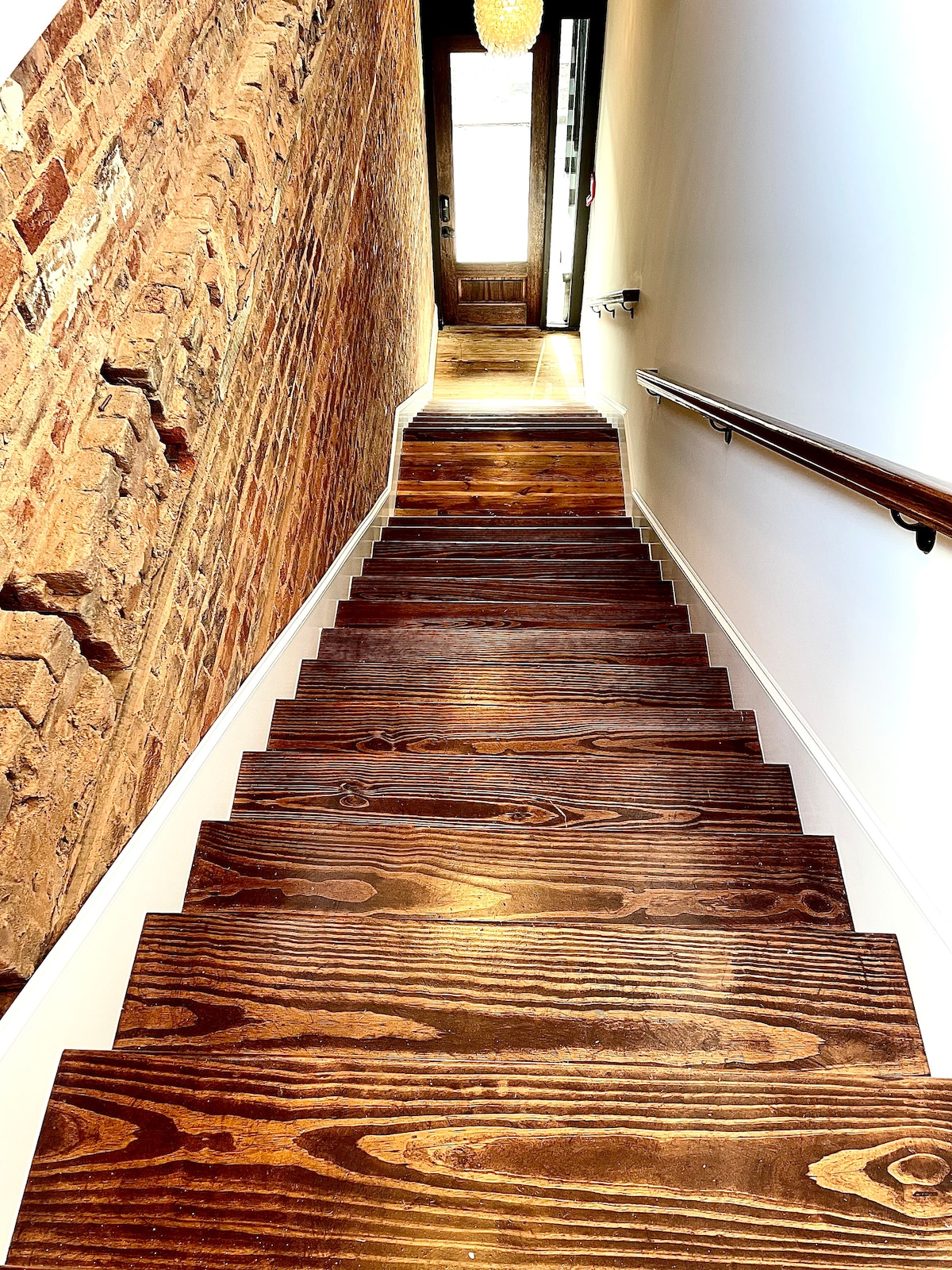 A wooden staircase is seen descending towards a bright entryway. The steps feature a rich, dark stain and are framed by exposed brick walls. A modern light fixture is visible at the top of the stairs, illuminating the path with natural light.
