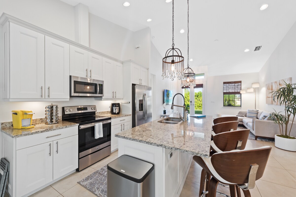 A modern kitchen is highlighted by white cabinetry and granite countertops. Stainless steel appliances are positioned for convenience, and bar stools are arranged at the spacious breakfast bar. Natural light filters in through large windows, illuminating the welcoming atmosphere.