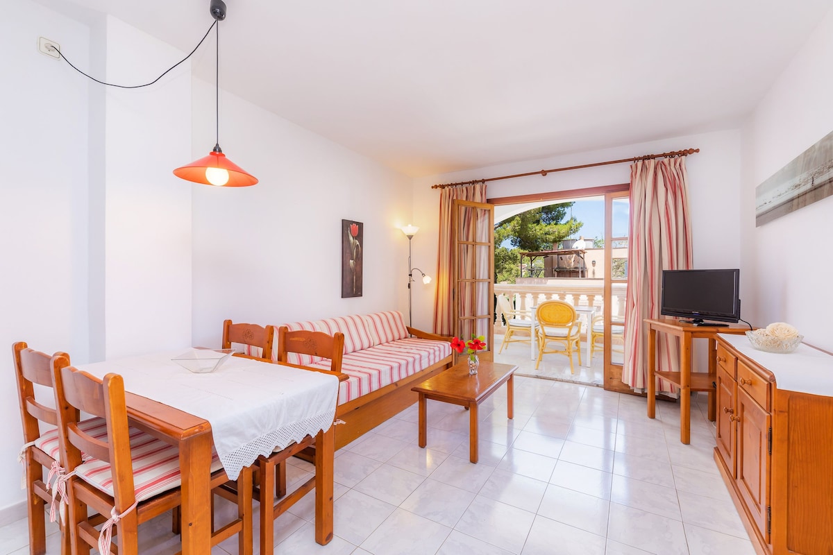 The spacious living and dining area features a sofa bed alongside a wooden table with four chairs. Natural light enters through a large window leading to a private terrace. A TV unit and decorative accents complete the inviting space.