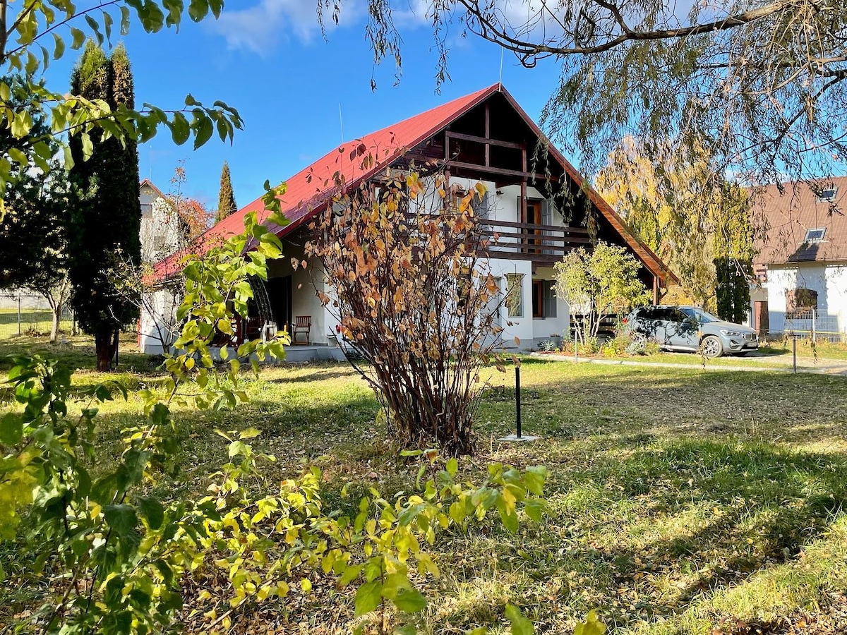 The exterior of Heritage Villa is showcased, revealing a two-story structure with a red roof and white walls. The villa is surrounded by a lush green garden, with trees and shrubs providing a natural backdrop. A parking area is visible at the side, accommodating a vehicle.