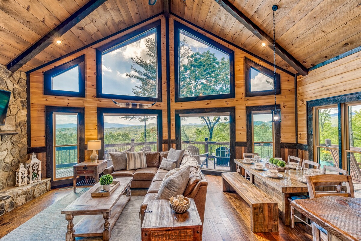 A spacious living area features large, angular windows that frame stunning mountain views. Natural wood accents and a stone fireplace create a warm ambiance. Comfortable seating is arranged around a rustic coffee table, and a dining area is visible, enhancing the inviting atmosphere.