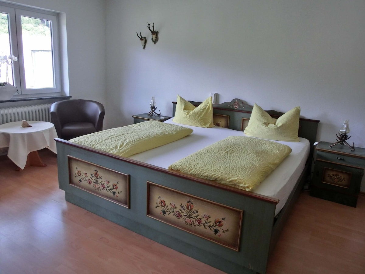 A spacious guest room features a double bed with two individual mattresses and decorative bed linens. Two lamps and a side table are positioned on either side of the bed. A seating area includes a chair and a small table, enhancing the room's cozy feel.