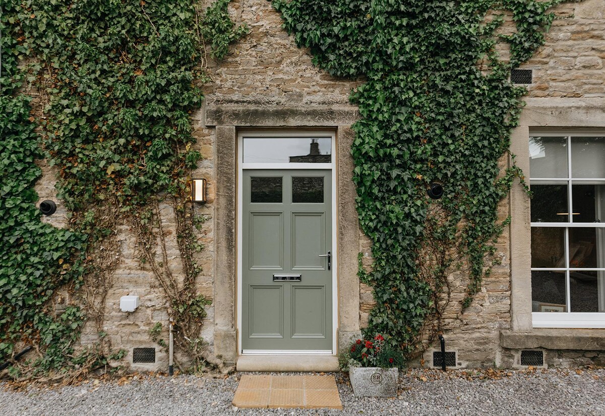 A charming exterior is displayed featuring a green door framed by a weathered stone wall. Lush ivy climbs the facade, and a decorative planter with flowers adds a touch of color near the entrance.