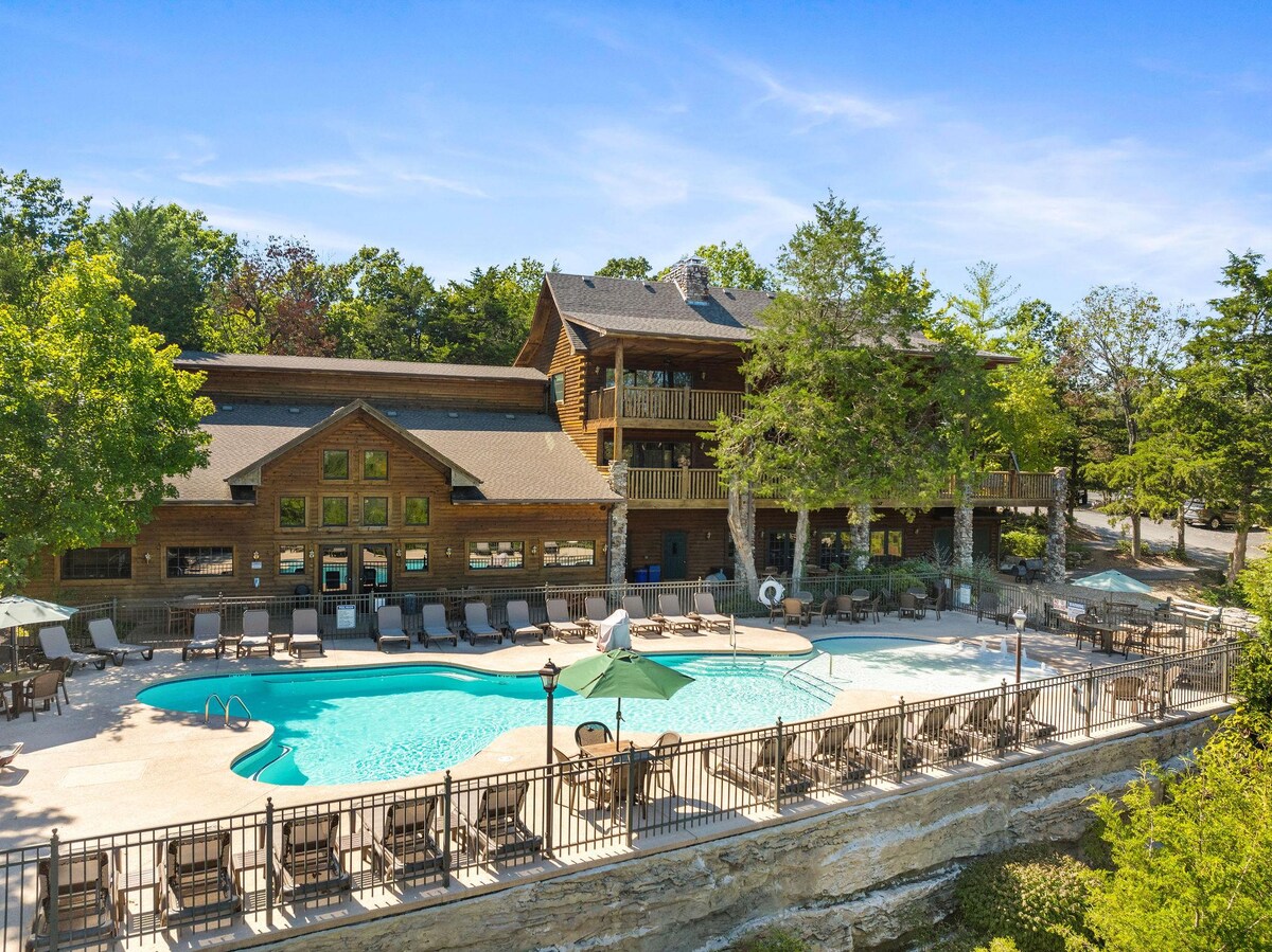 The image presents a spacious outdoor pool area surrounded by lounge chairs. A building with a wooden facade is in the background, and shaded umbrellas are positioned throughout the poolside. Lush green trees add a natural backdrop, contributing to a relaxing environment.