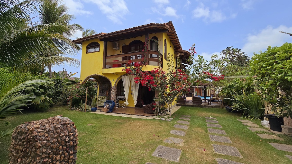A two-story yellow house surrounded by lush greenery. The exterior features a balcony adorned with floral accents and a pathway of stone leading through a manicured lawn. Sunlight filters through the leaves of palm trees, creating a serene outdoor atmosphere.