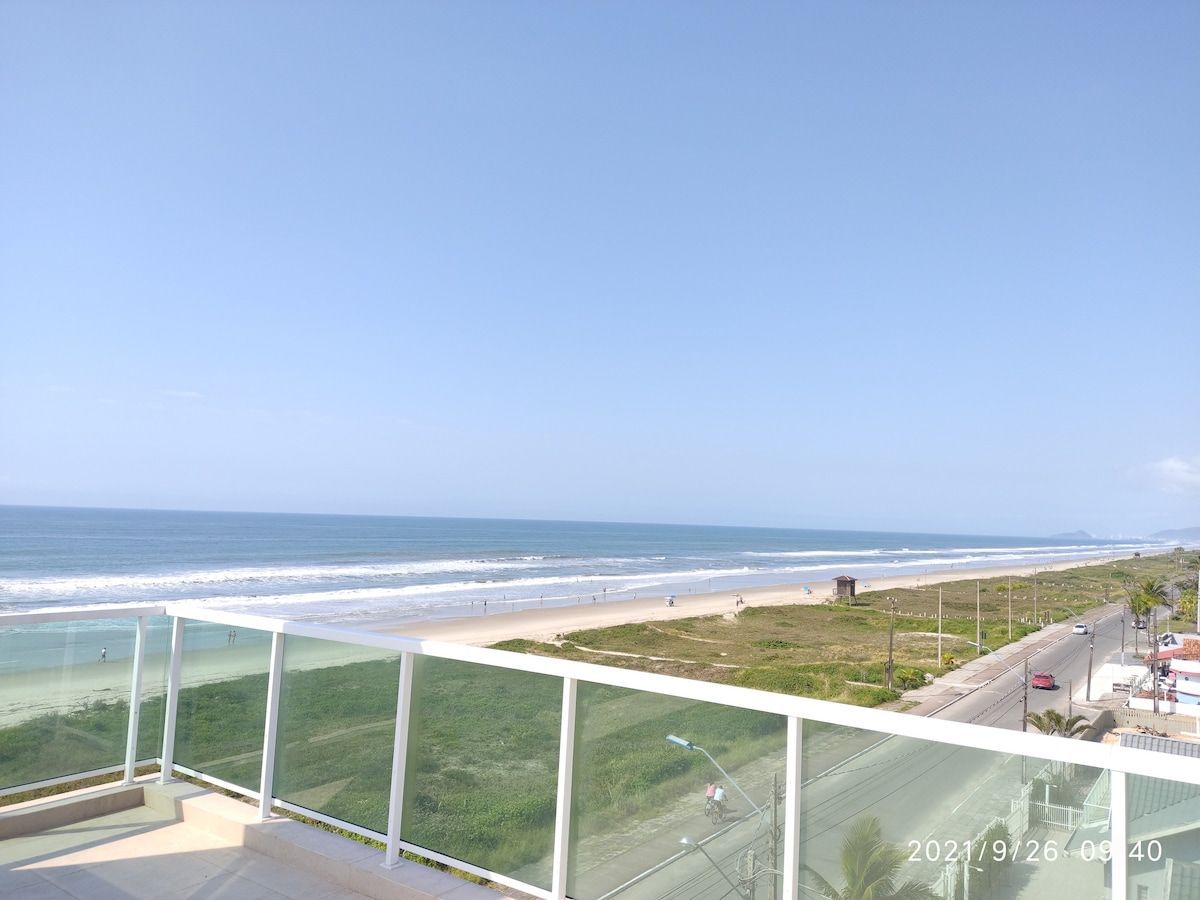 A panoramic view from a balcony captures the coastline, showcasing the sandy beach and calm waves meeting the shore. The ocean stretches across the horizon under a clear blue sky, with a glimpse of greenery and coastal road visible in the foreground.