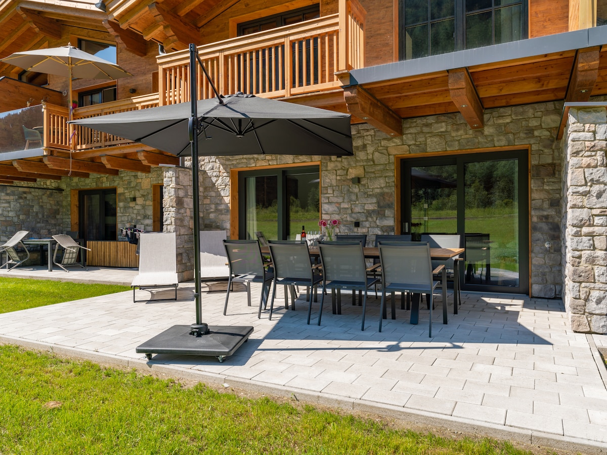 An outdoor patio features a dining set with multiple chairs and a large table, accompanied by a shaded umbrella. Sun loungers are positioned nearby on the green lawn. The natural stone facade of the building provides a rustic backdrop to the tranquil outdoor setting.