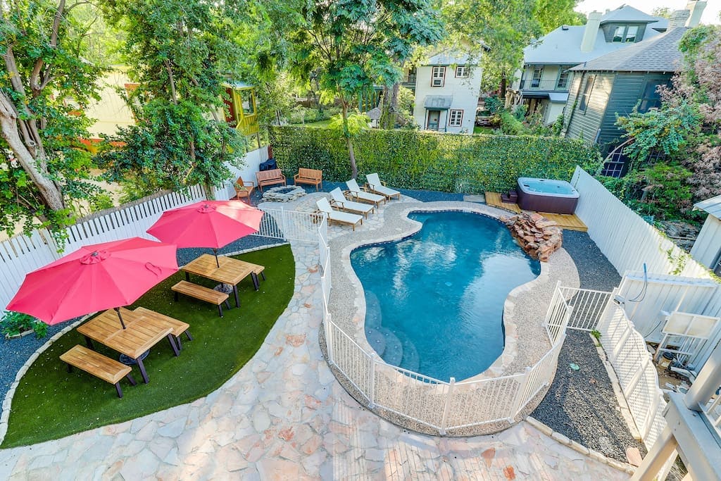A spacious backyard features a resort-style heated pool, surrounded by lounge chairs. Two red umbrellas provide shade over wooden picnic tables. A hot tub is positioned nearby, complemented by a rock water feature, all framed by lush greenery for privacy.