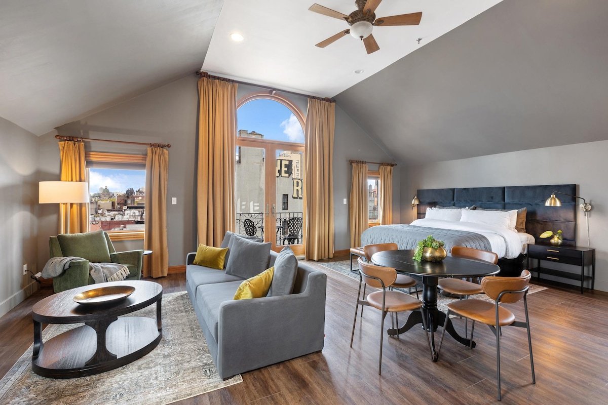 An open space features a king-size bed with crisp white linens, a cozy queen sofa bed, and a dedicated workspace. Large windows with gold curtains allow natural light to flood the room, highlighting a seating area with a green armchair and a round coffee table.