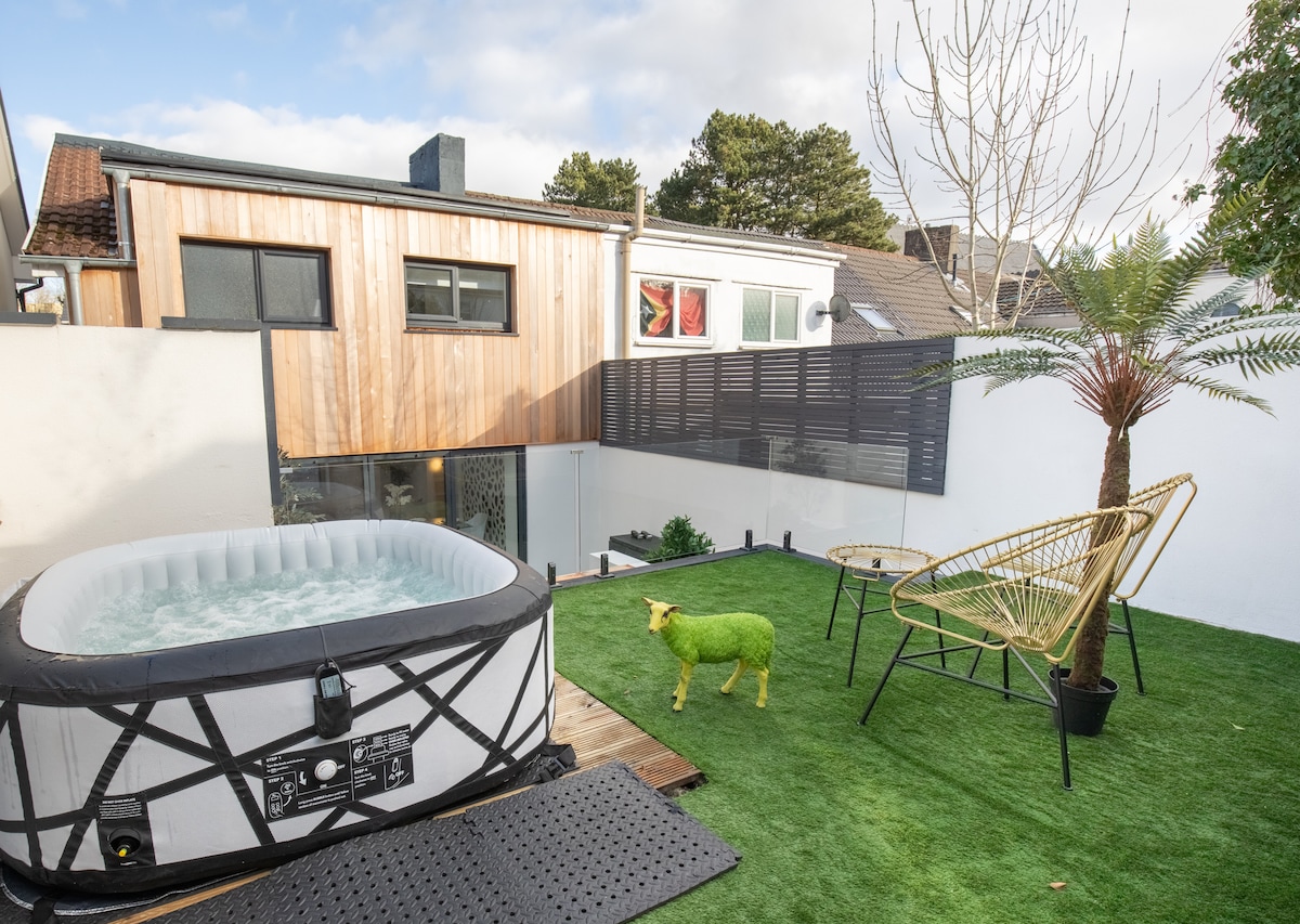 An outdoor area features a hot tub surrounded by green artificial grass. A decorative sheep sculpture adds a quirky touch, alongside a stylish chair and a palm tree. The background displays a contemporary wooden structure with large windows.