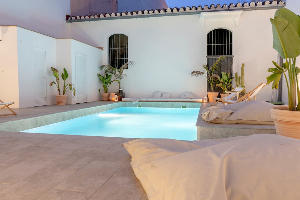 A serene pool area is highlighted, featuring a rectangular pool with illuminated water reflecting a calming blue. Surrounding the pool, potted plants add a touch of greenery, while comfortable loungers offer a relaxing space. The white walls create a bright and inviting atmosphere.