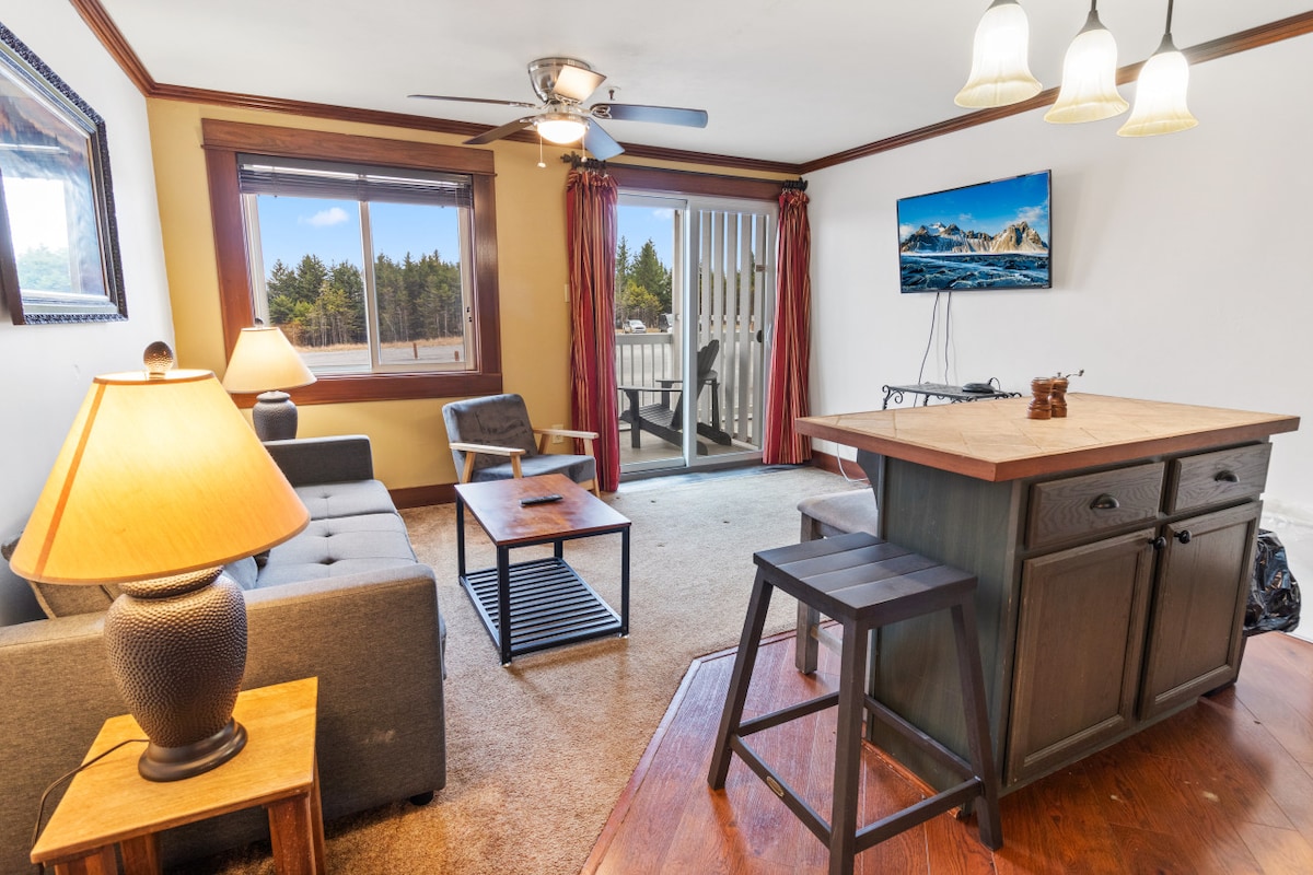 A living area is displayed with a comfortable gray couch and a wooden coffee table placed on a carpeted floor. Natural light enters through large windows adorned with curtains. A kitchen island, featuring a wooden top and stool, is visible alongside a wall-mounted flat-screen TV.