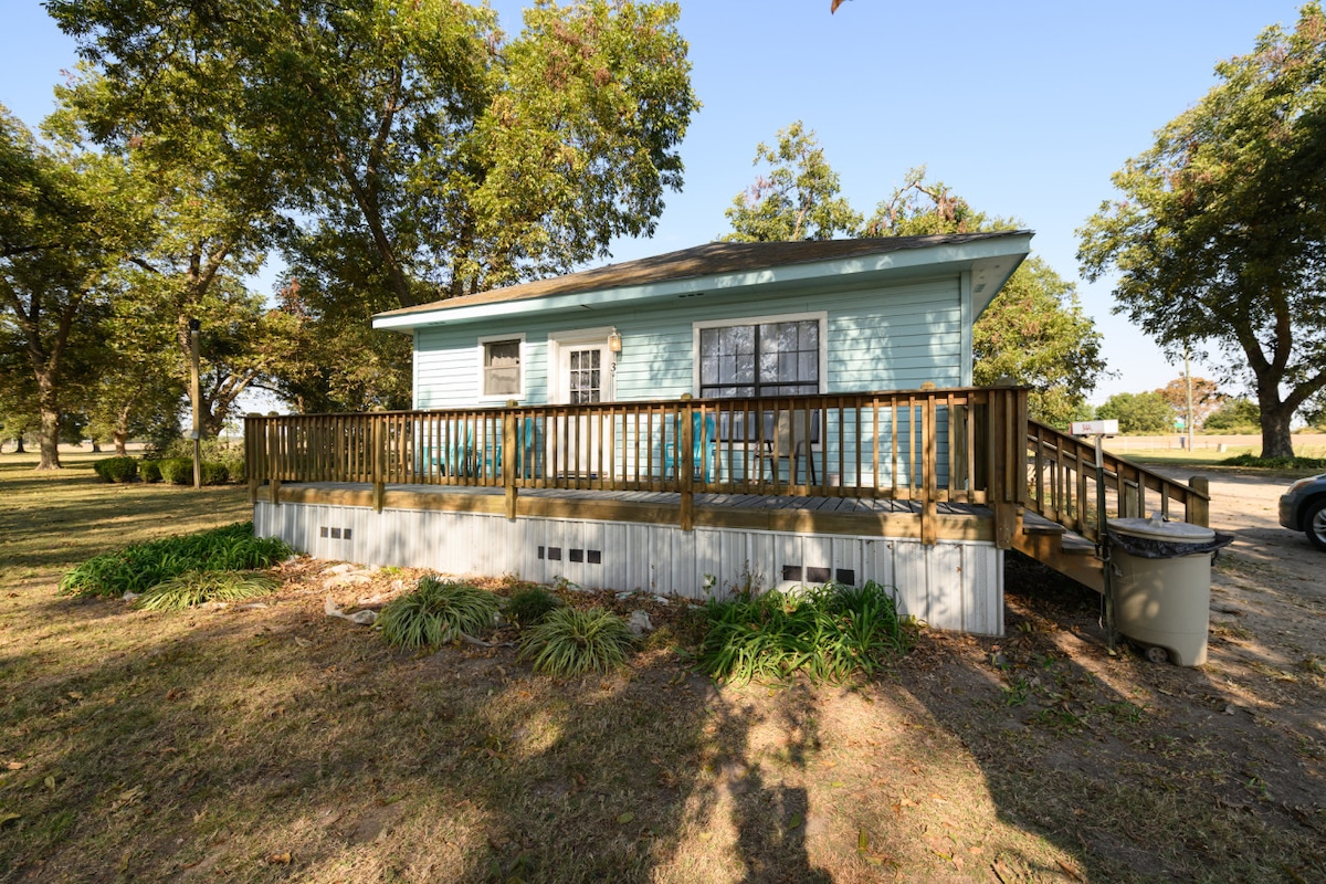 A charming cottage is set amidst grassy areas, featuring a wooden deck with seating. Large trees surround the building, providing shade. The exterior is painted in a soft blue hue with a window framed in white. A gravel pathway leads to the entrance.