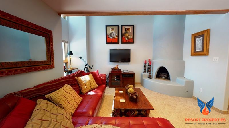 A welcoming living area features a red leather sectional sofa accented with decorative pillows. A wooden coffee table sits in front of the seating. A flat-screen TV is mounted on the wall, and natural light filters in through tall windows. A Kiva gas fireplace is positioned in the corner.