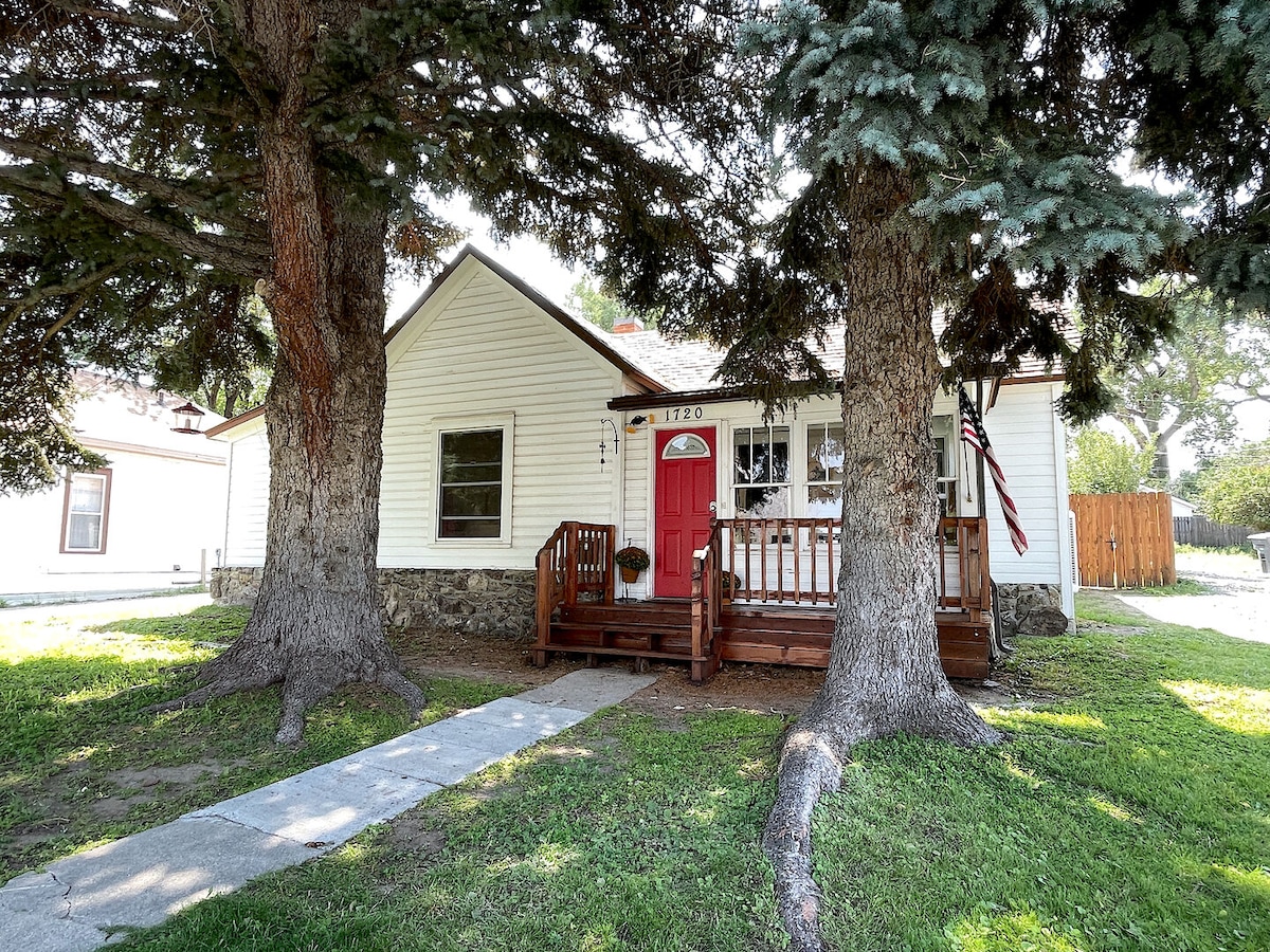 The charming exterior of the historic home features a welcoming red front door and a porch with wooden railing. Lush green grass surrounds the property, while tall trees provide shade. A pathway leads to the entrance, enhancing the inviting atmosphere of the home.