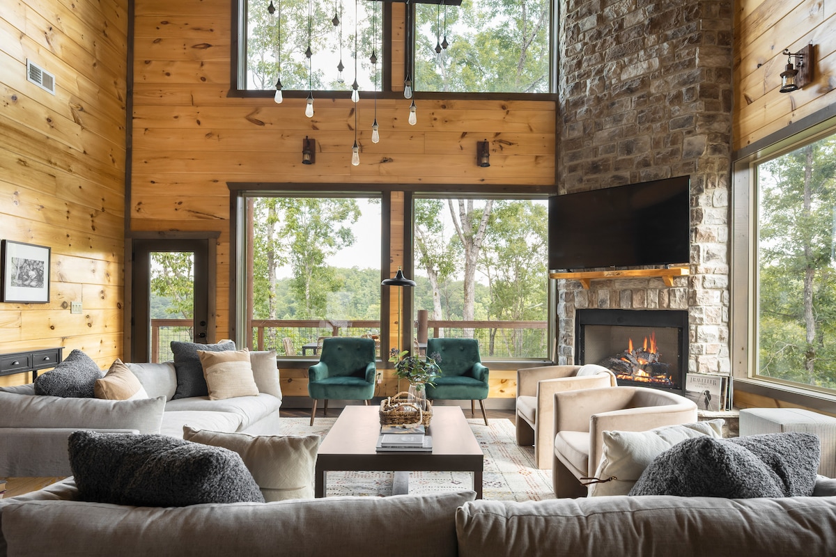 The spacious living area features large windows that provide panoramic views of the surrounding trees. A stone fireplace adds a cozy focal point, while comfortable seating arrangements consist of various sofas and armchairs adorned with cushions. Natural wood walls enhance the rustic atmosphere.
