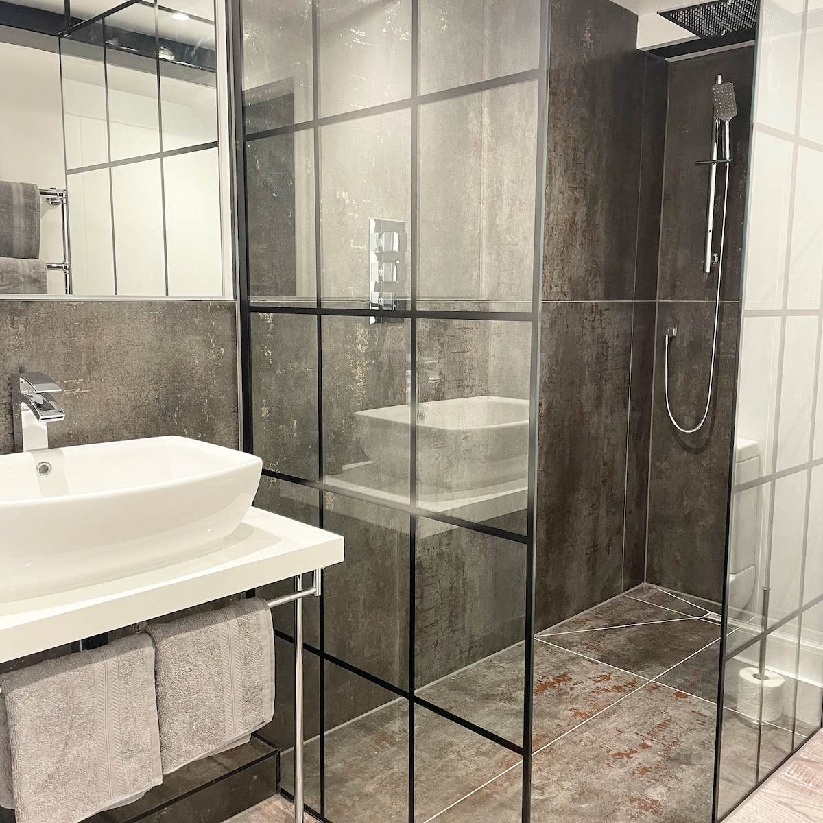 A modern bathroom features sleek glass partitions, a spacious shower with a rain showerhead, and a minimalist, floating sink basin. Towels are neatly arranged on a rack, complemented by contemporary tile flooring in muted tones.