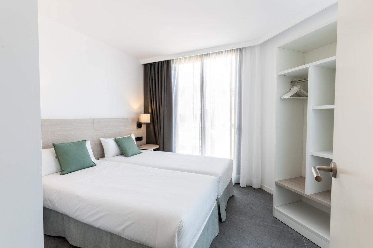 A cozy sleeping area features two spacious beds with white linens and green accent pillows. Natural light filters through large windows, while a minimalist closet with hangers provides storage. A neutral color palette contributes to a calm atmosphere.