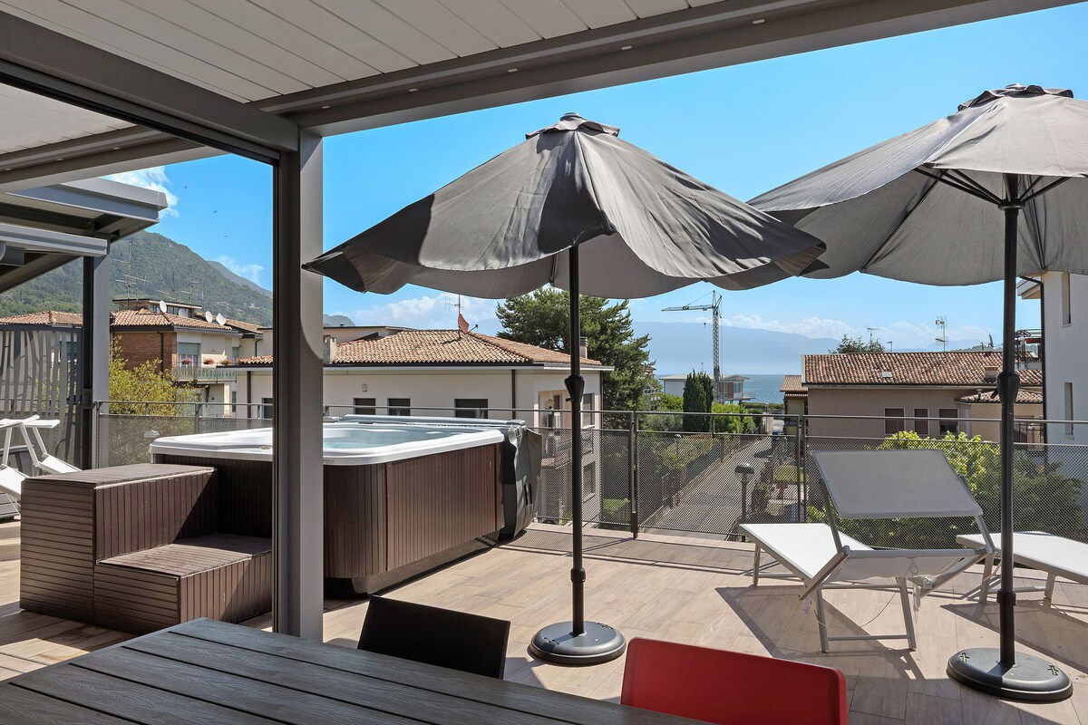 A well-furnished terrace features a dining table with colorful chairs, accompanied by two large umbrellas offering shade. A hot tub is positioned nearby, with lounge chairs arranged for relaxation. Scenic views of the surrounding landscapes and lake can be appreciated from this inviting outdoor space.