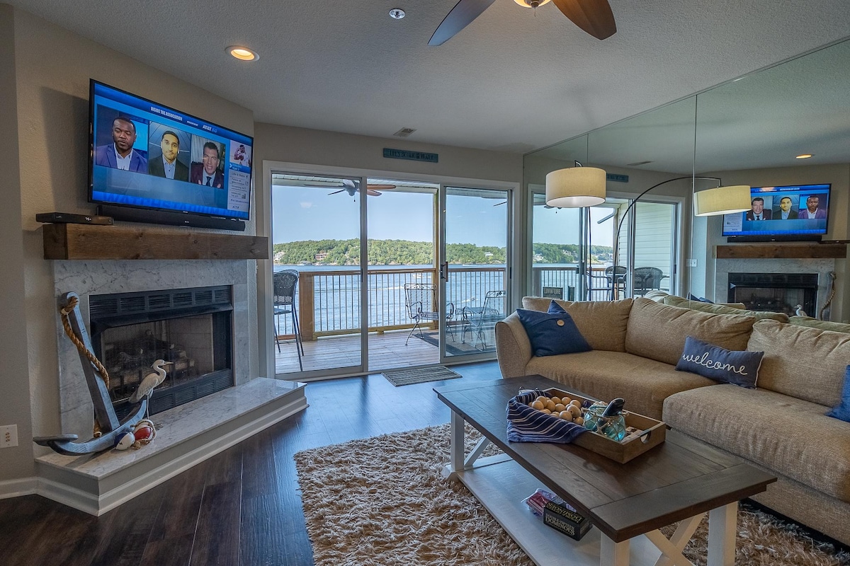 A cozy living area is depicted, featuring a soft sofa with decorative pillows and a coffee table adorned with a centerpiece. A fireplace is visible beside a wall-mounted television. Expansive glass doors provide a panoramic view of the lake and access to the balcony.