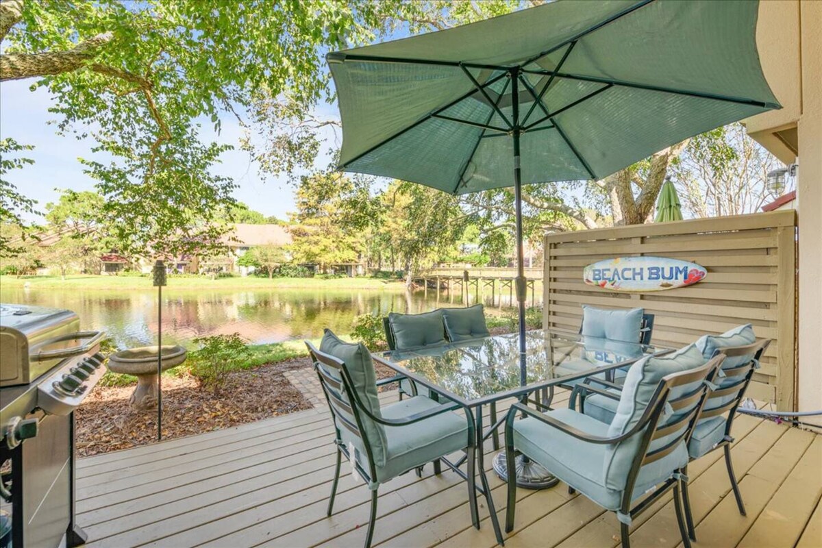 An outdoor patio area is presented with a glass dining table surrounded by six cushioned chairs. A large umbrella provides shade. A gas grill stands nearby, and scenic views of Horseshoe Lake are visible in the background, framed by lush greenery.