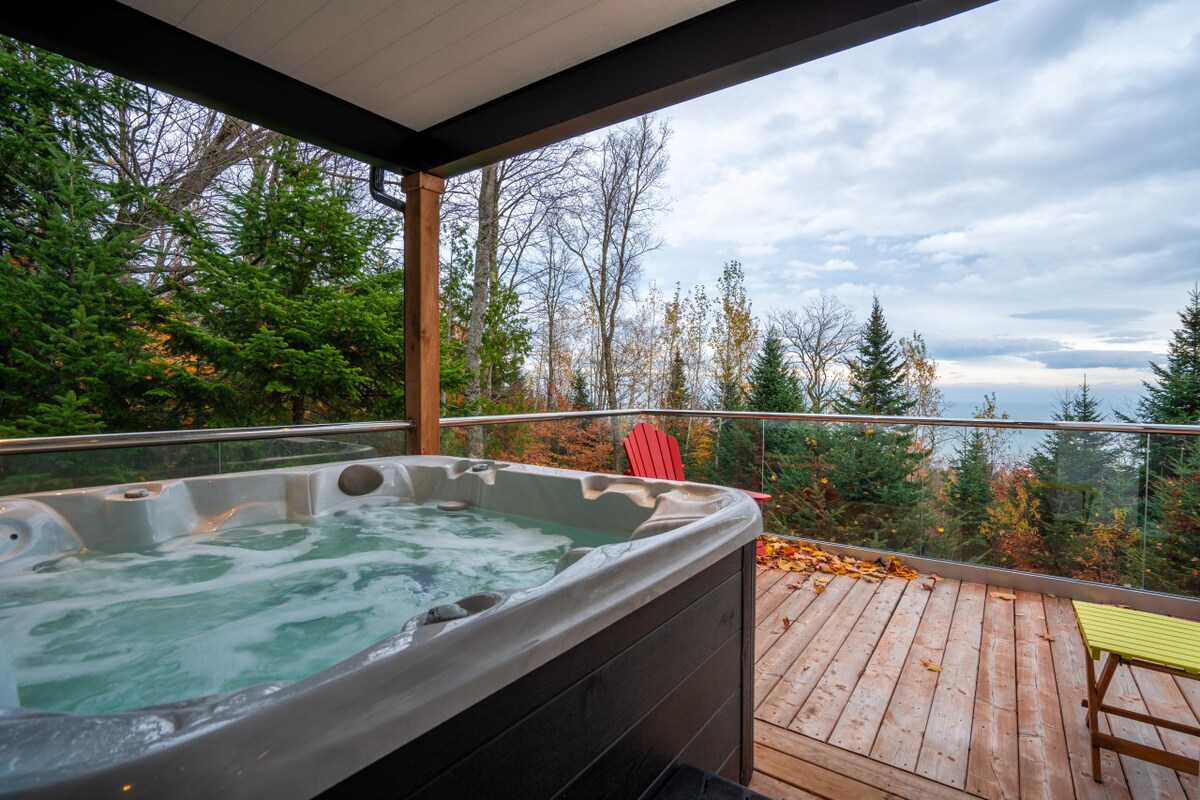 A private hot tub is situated on a spacious wooden deck, surrounded by trees displaying autumn foliage. The expansive view of the Saint Lawrence River is visible in the background, creating a peaceful outdoor retreat.