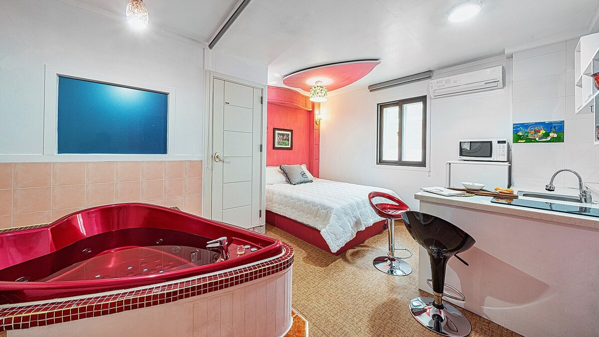 A room featuring a spacious bed positioned near a large window, allowing natural light to fill the space. A bright red spa tub adds a pop of color, while contemporary seating is arranged nearby. A compact kitchenette is visible, designed for convenience.