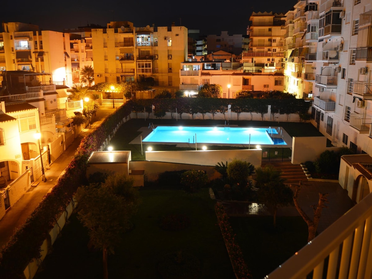 The communal pool is illuminated at night, surrounded by lush landscaping and softly glowing lights. Multiple residential buildings are visible in the background, contributing to the ambiance of the area. The pool measures approximately 10 by 20 meters, offering a relaxing retreat.