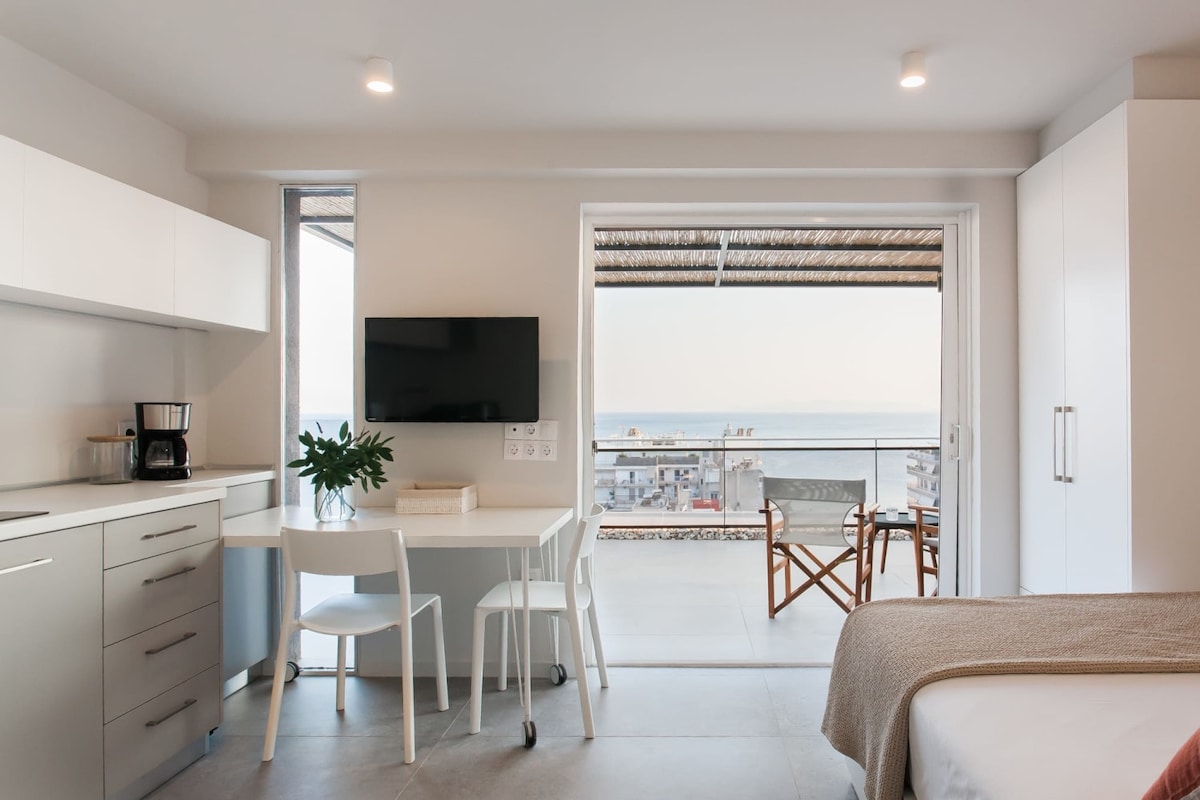 An open-plan studio showcases a functional kitchen area with built-in cabinets and a small dining table for two. A sliding glass door leads to a terrace with a sea view, while cozy director chairs provide additional seating. Natural light enhances the modern design.