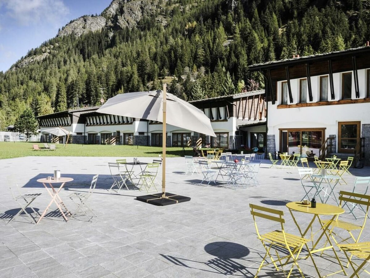An outdoor seating area features colorful metal tables and chairs arranged across a stone patio. A large canopy offers shade, while a backdrop of lush green trees and mountain slopes creates a tranquil setting. Bright sunlight enhances the inviting atmosphere.